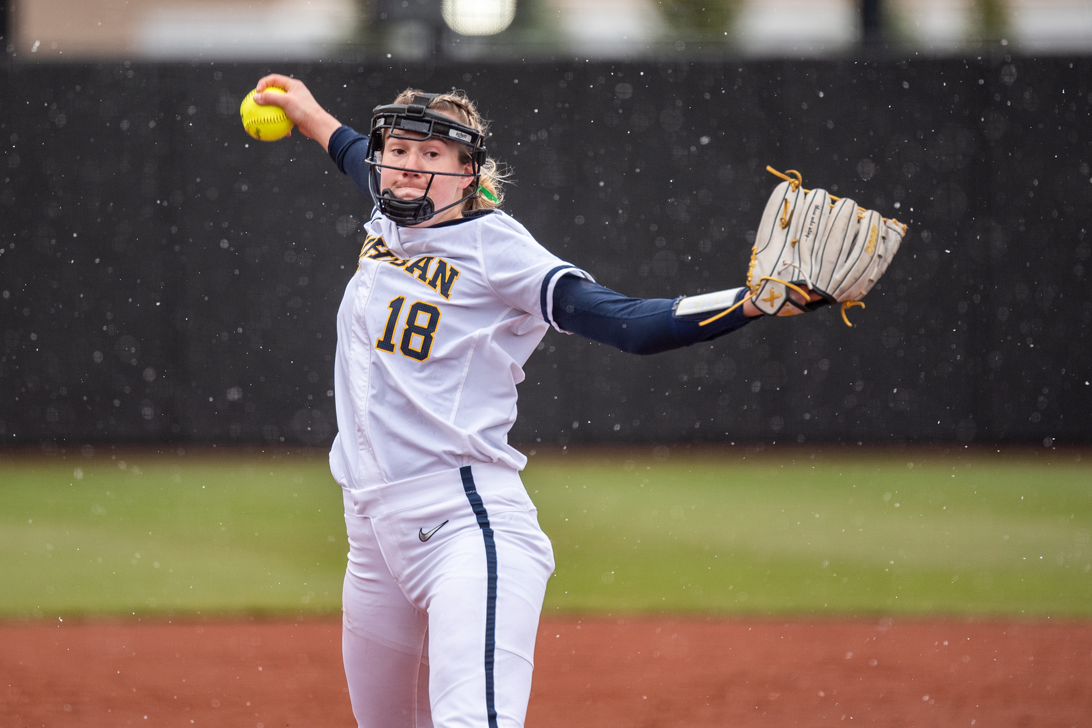Lauren Derkowski is wearing a white uniform and is mid pitch. Her hand with the mit is stretched out to the side while her other hand grips the ball as her arm swings. Behind her, snow is falling.